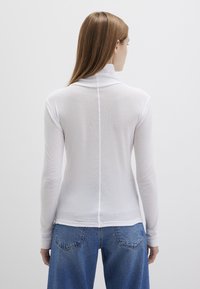 Weißes Langarmoberteil mit hohem Kragen, aus einem weichen, strukturierten Stoff und einer zentralen Naht auf der Rückseite. Kombiniert mit blauen Jeans.