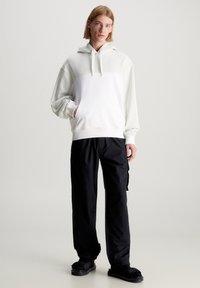 Sudadera blanca con un panel en gris claro que contrasta, que cuenta con un bolsillo tipo canguro, combinada con unos pantalones negros holgados y sandalias tipo slides.