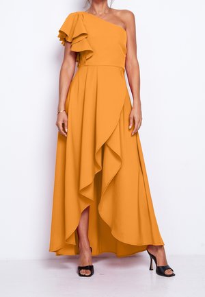 Femme portant une robe asymétrique jaune moutarde à épaules dénudées avec un détail volanté et des chaussures à talons hauts ouvertes noires, debout devant un fond blanc.