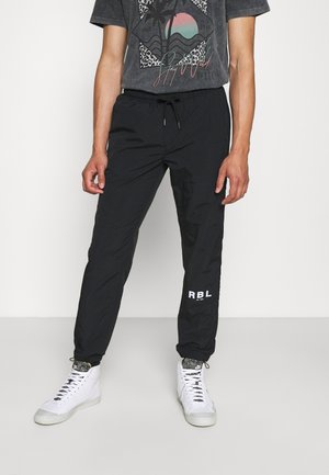 Person trägt schwarze Kordelzughose mit "RBL"-Schriftzug am linken Bein und weiße High-Top-Sneaker. Ein Teil eines grafischen T-Shirts ist sichtbar.