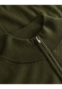 Olajzöld pulóver magas gallérral, fém cipzárral és sima, texturált kötött anyaggal. A nyakkivágás és a varrás részleteinek közeli képe.