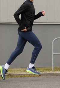 Persona che indossa un top a maniche lunghe nero, pantaloni da corsa blu navy, calzini bianchi e scarpe da corsa grigio-verdi che corre su un terreno pavimentato vicino a un muro.