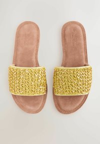 Sandalias planas con una plantilla de gamuza marrón y una tira tejida amarilla texturizada que cuenta con pequeños acentos en rosa y blanco. Diseño minimalista.