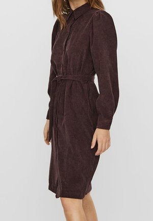 Vrouw draagt een knielange, donkerbruine corduroy jurk met lange mouwen, een kraag en een ceintuur in de taille.