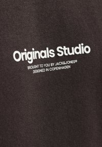 Zwarte katoenen stof met een gladde textuur bevat witte bedrukte tekst: "Originals Studio," "gebracht door JACK&JONES," en "ontworpen in KOPENHAGEN."