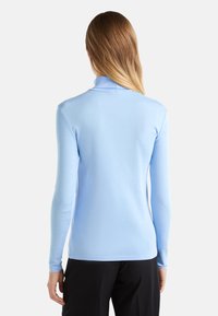 Kobieta z długimi blond włosami ubrana jest w jasnoniebieski golf i czarne spodnie, stoi tyłem na tle białego tła.