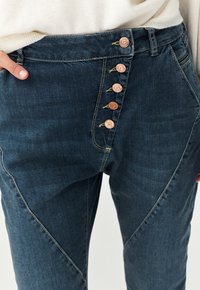 Dark denim trousers featuring a five-button closure, angled front seams, and contrasting yellow stitching. Smooth texture with a fitted design.