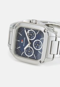 Kvadrāta formas sudraba metāla rokas pulkstenis ar zilu ciparnīcu, trīs papildus ciparnīcas, kas rāda dienu, datumu un 24 stundu laiku, un "Hilfiger" logo uz ciparnīcas.