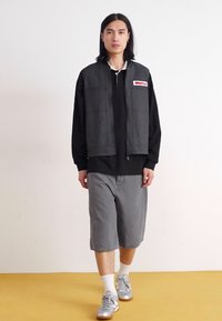Gray lightweight vest over a black long-sleeve shirt, paired with gray knee-length shorts and silver sneakers, featuring a front logo patch.