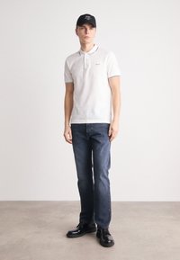Witte poloshirt met zwarte accenten, blauwe spijkerbroek en zwarte schoenen. Model draagt een zwarte pet met een logo. Glad textuur, casual pasvorm.