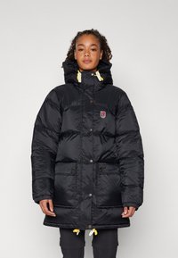 Black puffer jacket with a high collar, featuring yellow drawstring accents, snap buttons, and two front pockets. Made of glossy material.