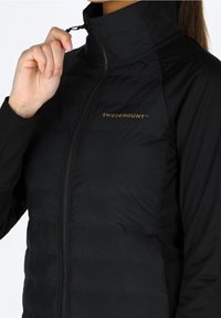 Veste noire isolante avec un col montant, dotée d'une fermeture éclair frontale et du logo "SWEDEMOUNT" en or sur la poitrine. Tissu lisse et léger.