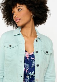 Hele sinine teksajakk, millel on nööpidega esitasku, kaks rinna-taskut ja hõbedane riistvara, kantud lillemustrilise kleidi peal.