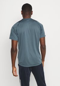 Man med blå atletisk t-shirt och mörka träningsbyxor som står med ryggen mot kameran mot en ren vit bakgrund.