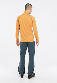 Langärmlige orange Basis-Schicht mit hohem Kragen und grauen Nähten, kombiniert mit blauen Skihosen mit seitlichen Reißverschlüssen und schwarzen Akzenten.