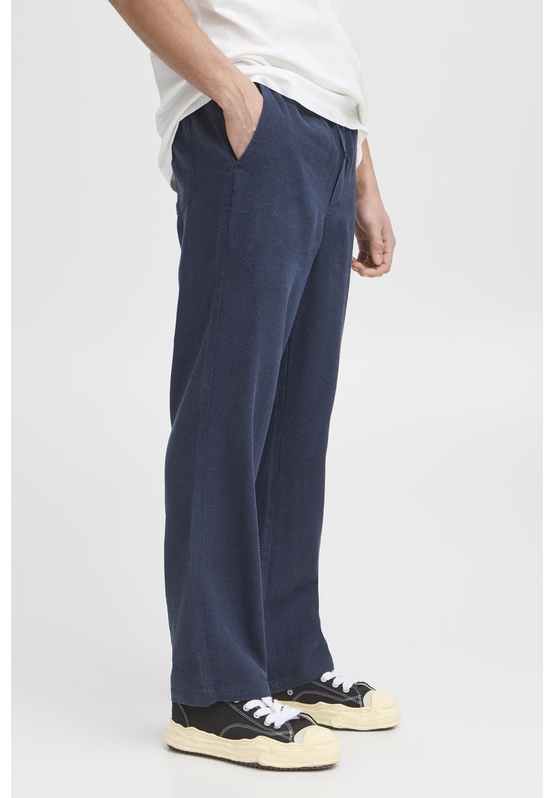 Navy blue linen trousers with a relaxed fit, featuring side pockets and a smooth texture. Paired with black sneakers and white laces.