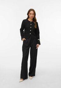 Crna blejzerica s zlatnim gumbima, uparena s crnim širokim hlačama. Glatki materijal, kroj koji pristaje tijelu i bež visoke pete zašiljenog vrha upotpunjuju odjevnu kombinaciju.