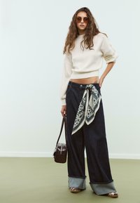 Pantaloni in denim a gamba larga con orlo in denim a contrasto, accessoriati con una sciarpa a fantasia legata in vita e una borsa a spalla marrone. Maglione cropped color crema.