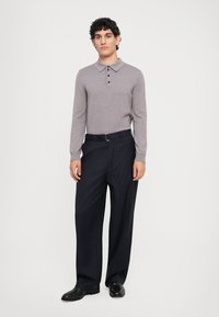 Pelēka garām piedurknēm polo krekls ar trīs pogām, kopā ar tumši zilu svītrainu biksēm. Modelis valkā melnas kurpes, stāvot uz neitrāla fona.