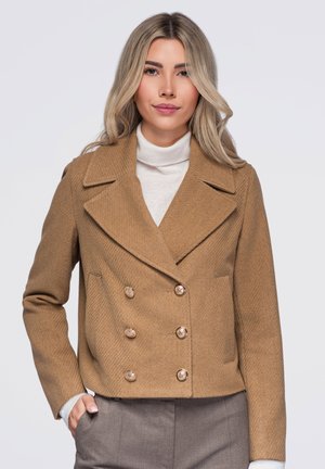 Femme aux cheveux blonds portant un manteau croisé beige sur un col roulé blanc et un pantalon gris, debout avec une main dans la poche.