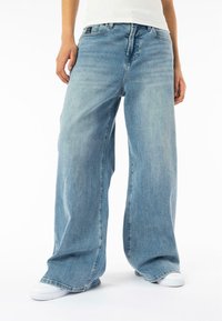 Jean large bleu clair en denim, avec une fermeture à bouton sur le devant et des poches. Des chaussures en toile visibles en bas.