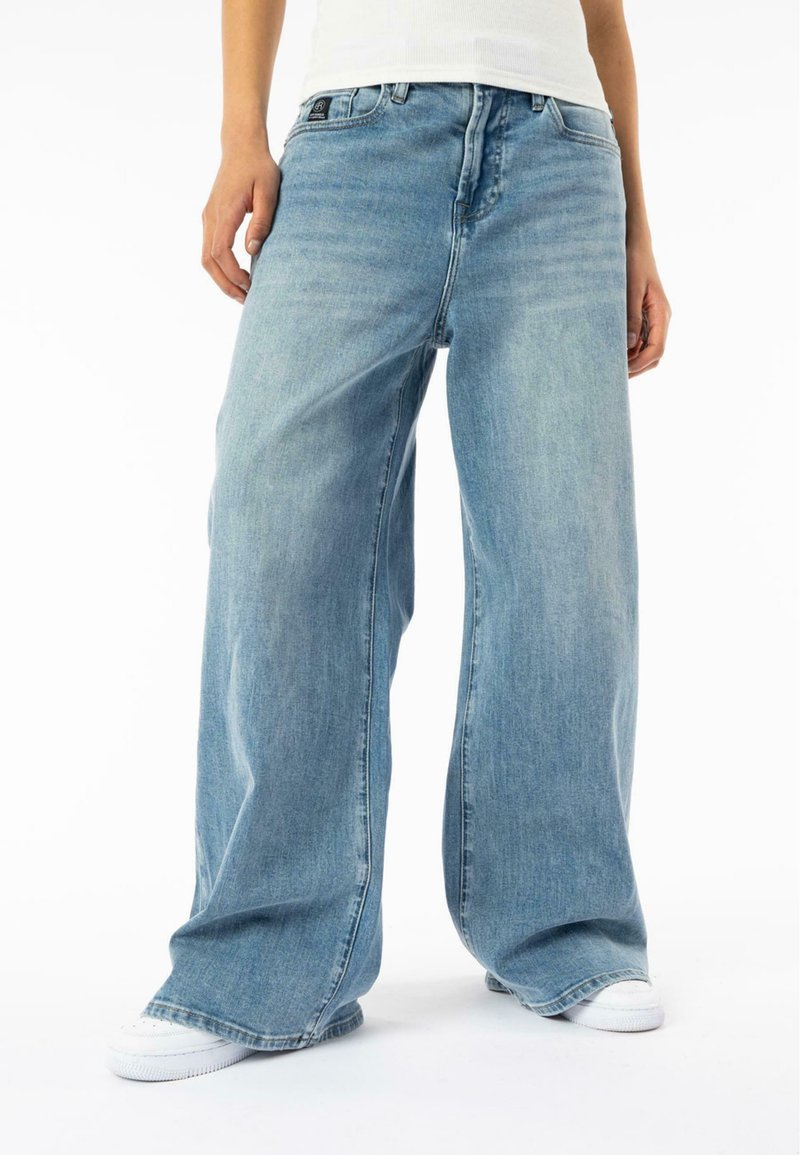 Jean large bleu clair en denim, avec une fermeture à bouton sur le devant et des poches. Des chaussures en toile visibles en bas.
