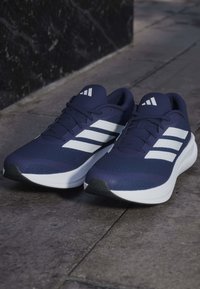 Ζευγάρι μπλε και άσπρα αθλητικά παπούτσια Adidas τοποθετημένα σε πέτρινο πάτωμα κοντά σε σκοτεινό τοίχο.
