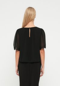 Γυναίκα με μακριά ξανθά μαλλιά που φοράει μαύρο μπλουζάκι με διαφανείς κοντές μανσέτες και μικρή κουμπωτή τρύπα στο πίσω μέρος του λαιμού.