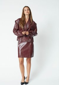 Giacca in pelle sintetica bordeaux con spalle oversized e polsini arricciati, abbinata a una gonna a tubino coordinata, entrambe con una finitura lucida.