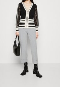 Vrouw draagt een zwart-wit gestreepte cardigan met kanten mouwen, lichtgrijze cropped broek, zwarte laarzen met hak en draagt een zwarte handtas.