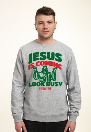 Šedá mikina s tučným textem: "JESUS IS COMING LOOK BUSY" v zelené a červené barvě. Design zahrnuje grafiku postavy s vztyčenýma rukama.