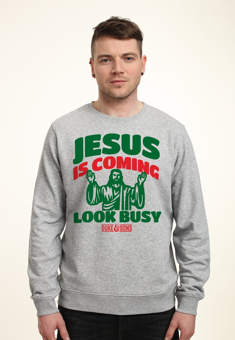 Šedá mikina s tučným textem: "JESUS IS COMING LOOK BUSY" v zelené a červené barvě. Design zahrnuje grafiku postavy s vztyčenýma rukama.