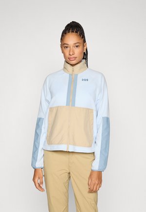 Gaiši zila un bēša rāvējslēdzēja jaka ar augstu apkakli, kontrastējošiem paneļiem un zilu logo uz krūtīm. Gluda tekstūra un sportisks dizains.