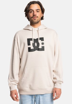 Ljus beige hoodie i mjukt tyg, med en stor svart DC-logotyp i mitten, en framficka och justerbara dragsko på huvan.