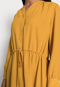 Vestido de color mostaza hecho de una tela ligera, con escote en V, mangas largas, cintura elástica con cordón y sutiles patrones de puntos blancos.