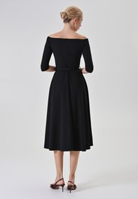 Schwarzes schulterfreies Kleid mit drei Viertelärmel, tailliertem Oberteil, gerafftem Bund, fließendem Rock und Reißverschlussdetails auf der Rückseite.