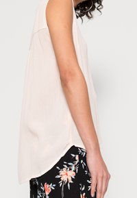 Mujer vestida con blusa sin mangas de color rosa claro y pantalones negros con un patrón floral naranja y azul, mostrada de lado con el brazo relajado.