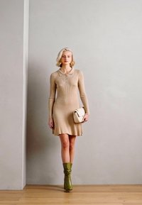 Knit beige pitkähihaisella mekolla, jossa on kaulus, nappikoristeet ja väljät helmat; yhdistettyinä vihreisiin korkokengtiin ja valkoiseen käsilaukkuun.