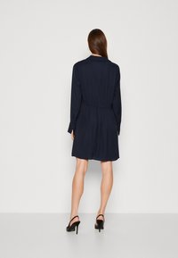 Navyblaues Kleid mit Kragen, elastischem Bund, langen Ärmeln und fließender Silhouette. Getragen mit schwarzen High Heels.