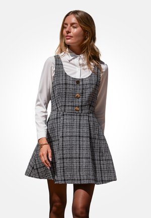 Frau trägt ein ärmelloses schwarz-weiß kariertes Kleid mit großen Knöpfen über einem langärmligen weißen Hemd mit Kragen, steht vor einem weißen Hintergrund.