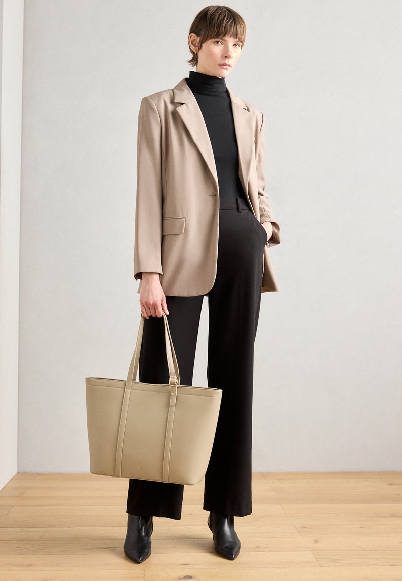 Bolsa tote beige con asas dobles, hecha de un material suave, que presenta un diseño minimalista y sutiles acentos de costura, sostenida por un modelo.