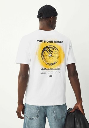 Bílý bavlněný tričko s žlutým kruhovým grafickým prvkem a černým textem na zádech, uvádějící koncertní data a místa pro "The Stone Roses."