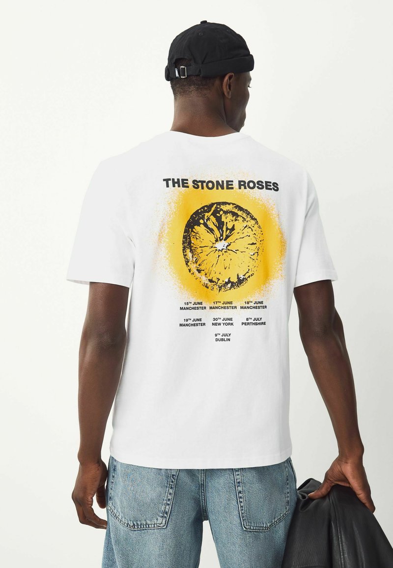 Valkoinen puuvillapaita, jossa on keltainen ympyräkuvio ja musta teksti selässä, jossa on luettelo konserttipäivistä ja -paikoista "The Stone Roses" -yhtyeelle.