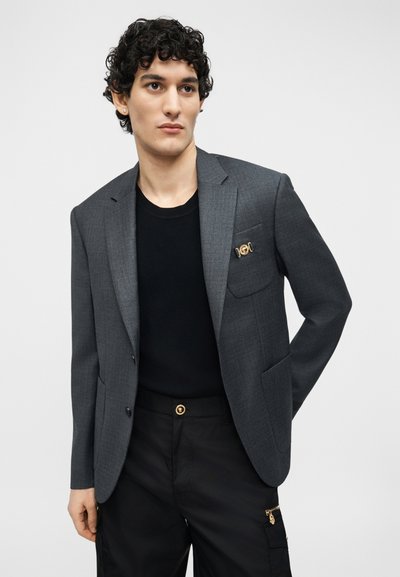 Homme aux cheveux bouclés portant un blazer gris foncé avec une épingle dorée, une chemise noire et un pantalon noir avec une poche à bouton doré, sur fond blanc.