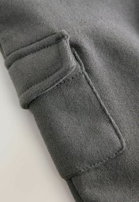 Szary materiał o gładkiej fakturze i widocznym szwie. Posiada naszywaną kieszeń zintegrowaną z projektem dla zwiększenia funkcjonalności.