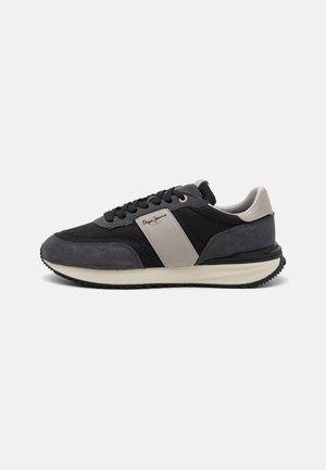 Zapatillas negras y grises con una parte superior de ante y malla, que presentan una banda beige prominente y una suela texturizada para mayor tracción.