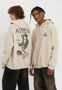Beige hoodie with front pouch pocket. Back features black graphic of a figure and text "ATHENS." Fabric appears soft with a relaxed fit.