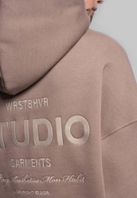 Ylihankkinen huppari vaaleanruskeasta kankaasta, jossa on kohotettua valkoista brodeerausta "STUDIO" ja "WRSTBVR" selässä sekä teksturoitua materiaalia.