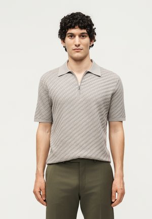 Homme aux cheveux noirs bouclés portant un polo beige texturé à manches courtes et un pantalon vert olive, debout devant un fond clair uni.