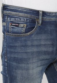 Tmavomodré džínsy z denimu s klasickým dizajnom s piatimi vreckami, viditeľným prešívaním a malou nášivkou s logom na prednom vrecku.